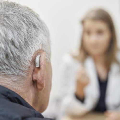 Close-up of a senior man with hearing aid on his ear visit his doctor at hearing clinic. Over the shoulder view of a elderly man wearing hearing machine sitting at medical clinic and listening to the female doctor advice.